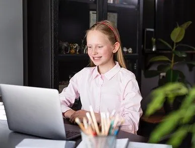 Smiling young girl wearing a pink shirt and headband sitting at a desk using a laptop in a room with a black cabinet and plants.