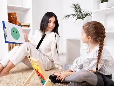 Woman showing a clipboard with a green smiley face to a girl sitting on a carpeted floor in a bright room with toys and books.