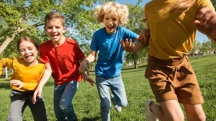 Four children holding hands and running on green grass in a sunny park.