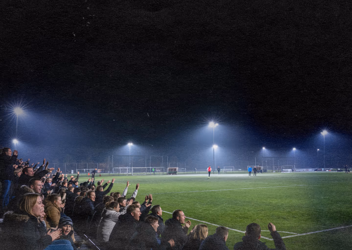 Crowd of spectators cheering and clapping at a nighttime soccer match under bright stadium lights.