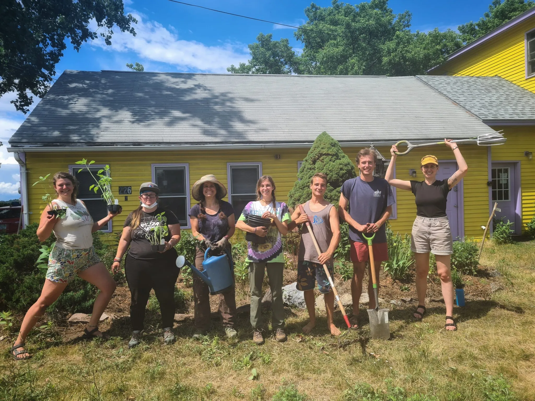 Group of people standing in front of a yellow house holding gardening tools and plants.