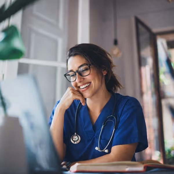Smiling female healthcare professional in blue scrubs and glasses using a laptop indoors.