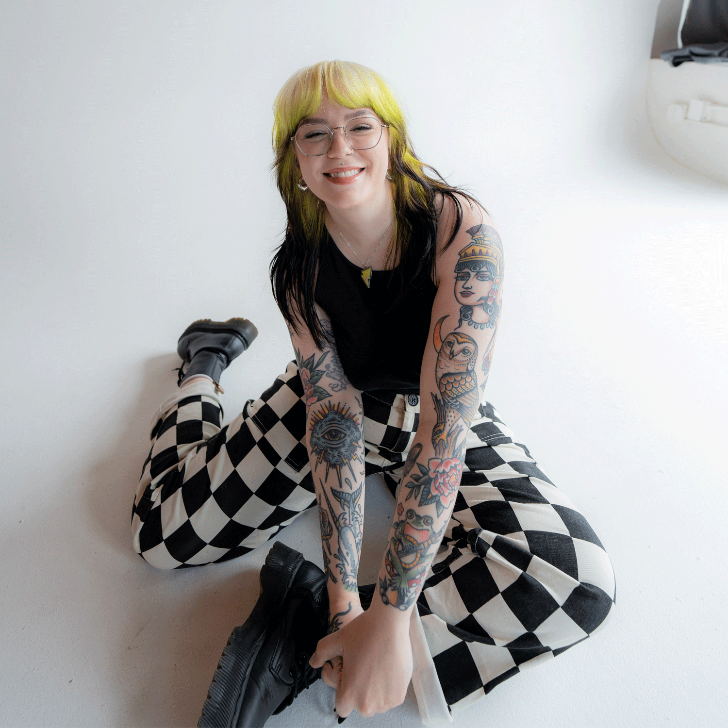 Marci, brand and web designer, is smilinig and has green and black hair, glasses, and tattoos on arms wearing black top, checkered pants, and black boots sitting on the floor.
