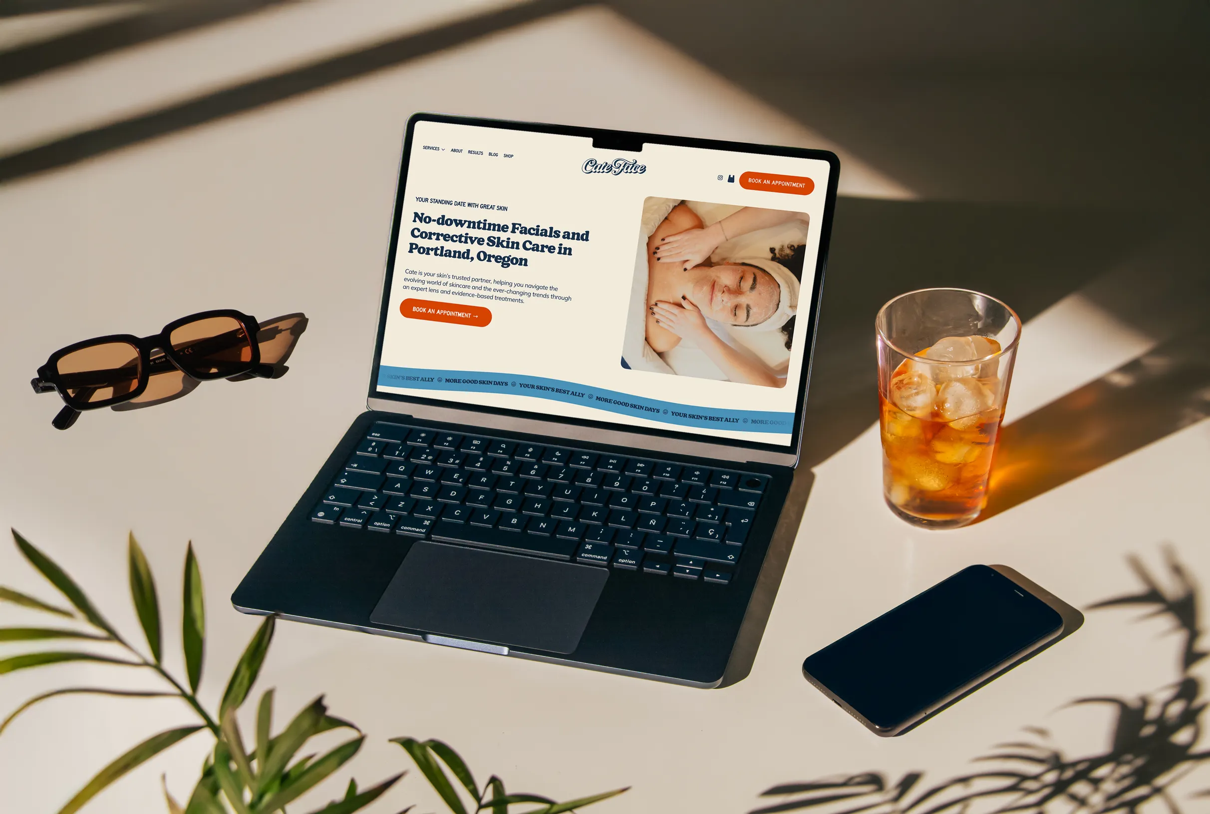 Laptop on a table displaying a skincare website with a facial treatment promotion, next to sunglasses, a glass of iced tea, and a smartphone.
