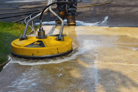 Person using a yellow surface cleaner to power wash a concrete sidewalk next to grass.