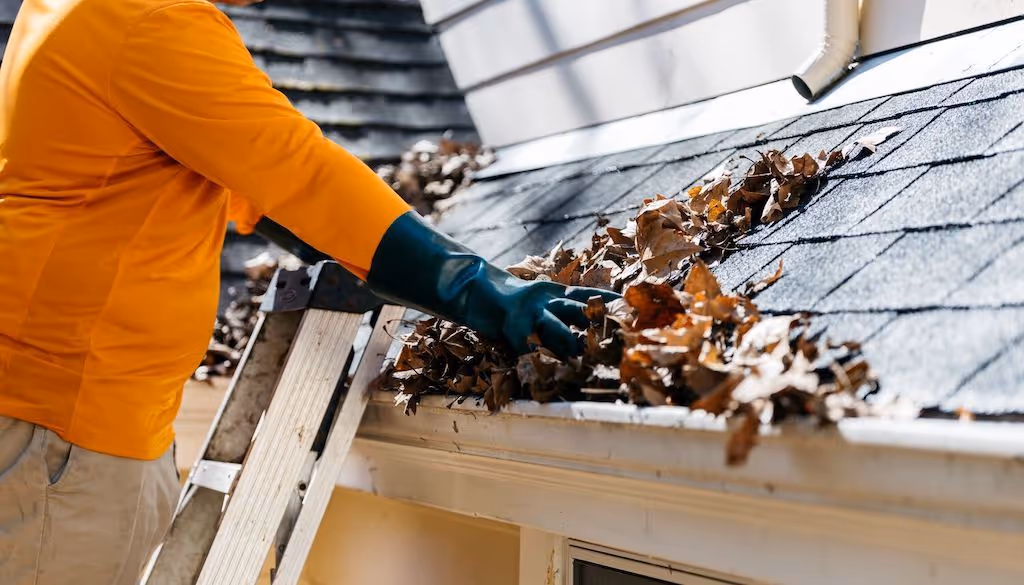 Person wearing orange shirt and green gloves cleaning leaves from a rooftop gutter while standing on a ladder.
