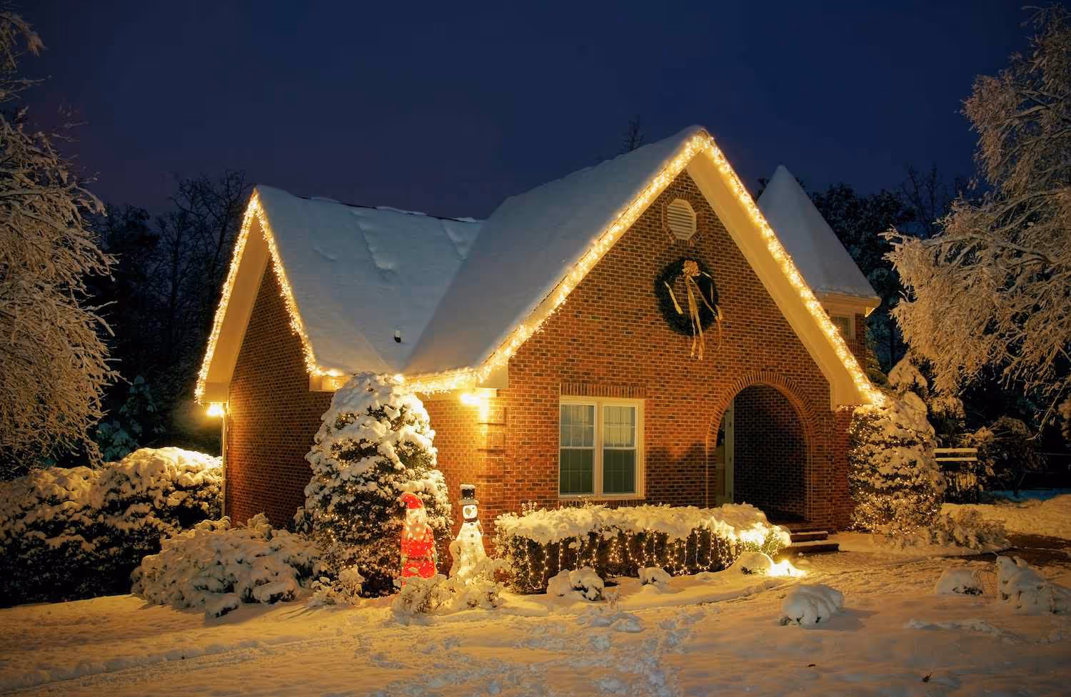 Snow-covered brick house decorated with Christmas lights, wreath, and illuminated holiday figures at night.