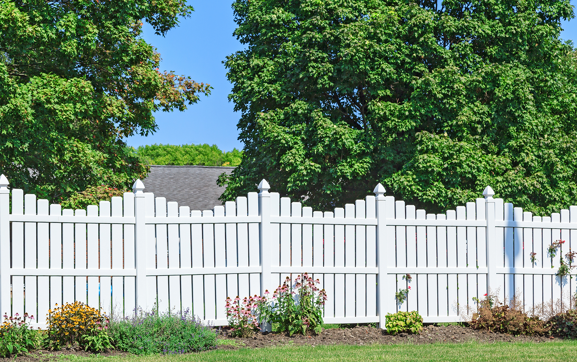 White picket fence with decorative tops in a garden with flowers and large green trees in the background under a clear blue sky.