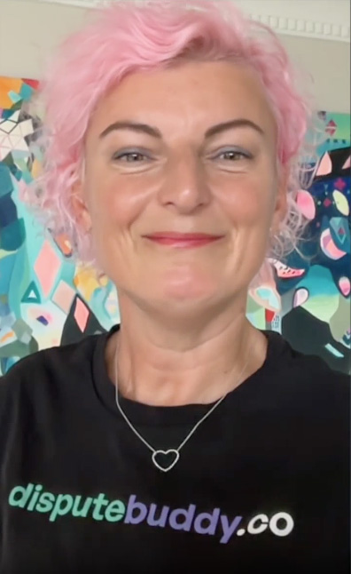Jenny Rudd Smiling with curly pink hair wearing a black shirt with 'disputebuddy.co' text and a heart-shaped necklace, colorful abstract painting in the background.