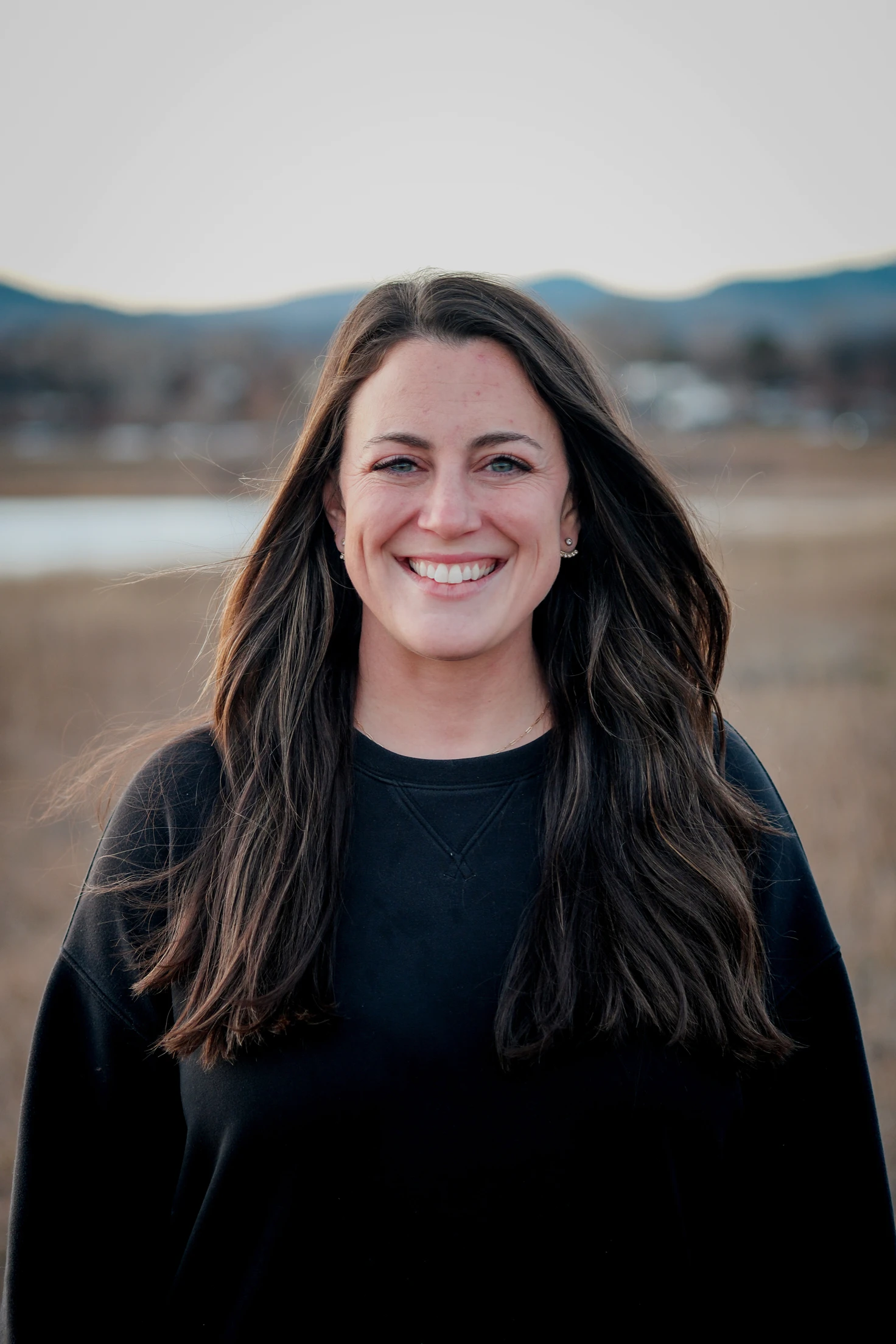 Smiling woman with long dark hair wearing a black sweater outdoors with mountains and water in the background.