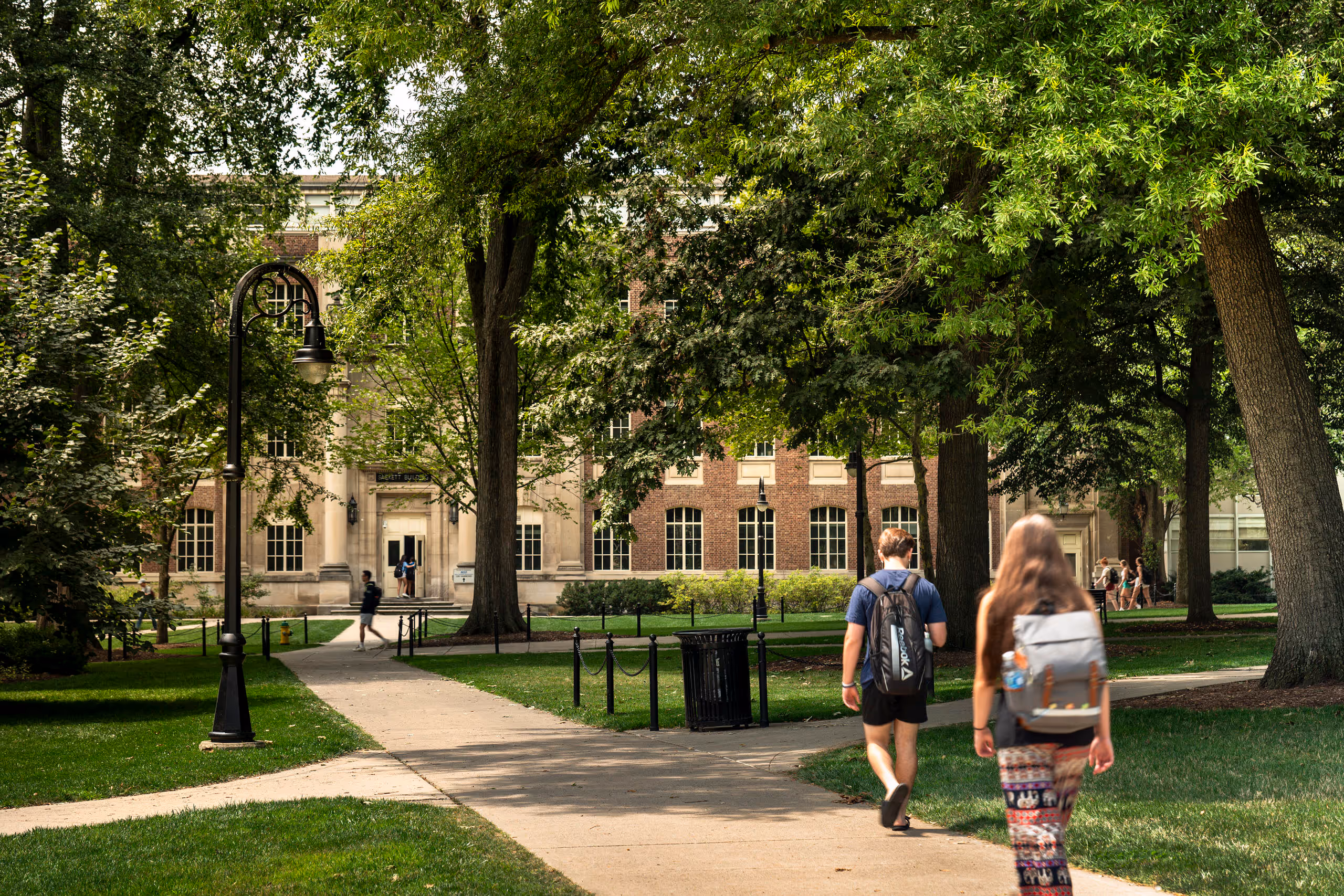 Students walking on a tree-lined path toward a large academic building on a green university campus.