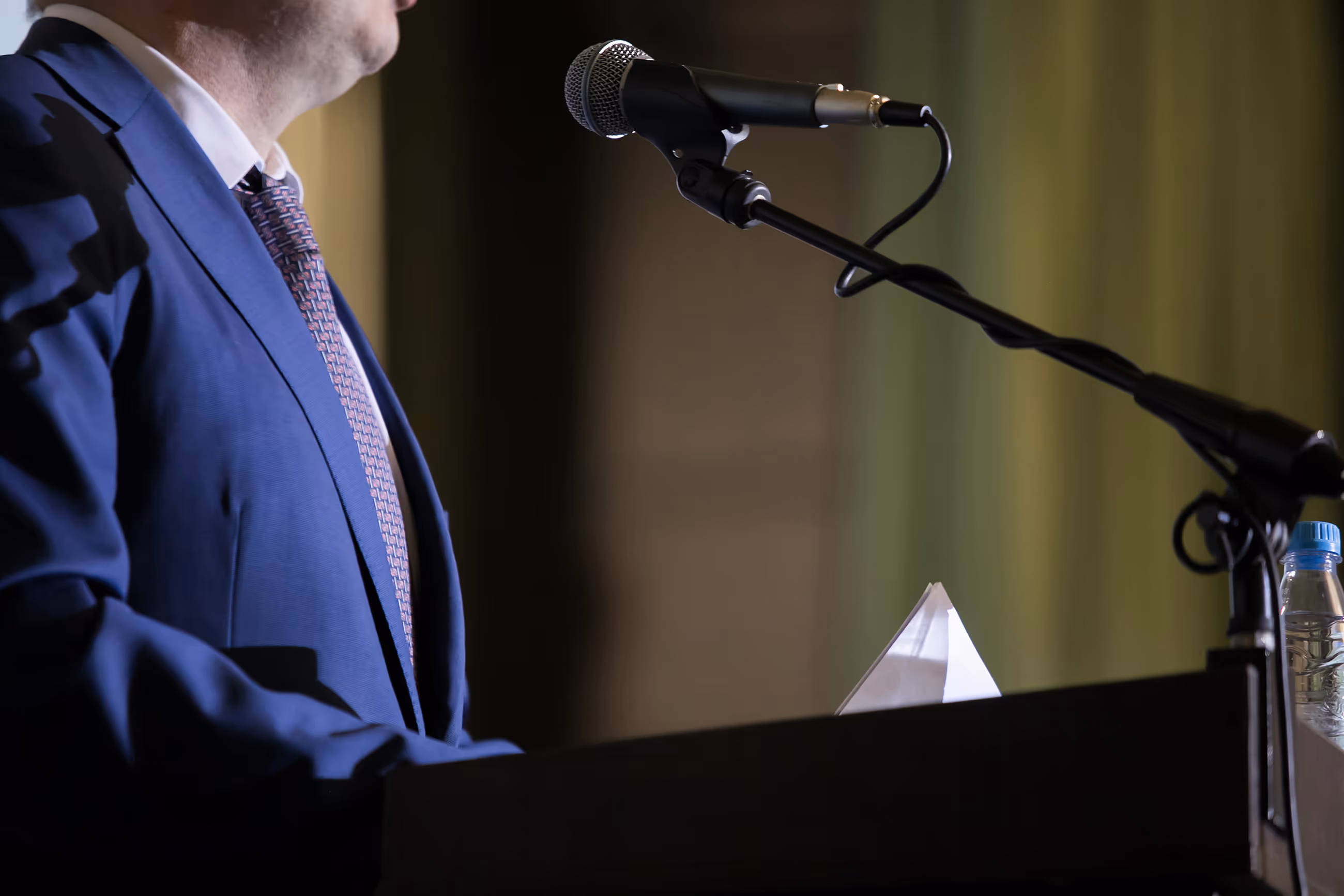 Man in blue suit and patterned tie speaking at podium with microphone and water bottle.