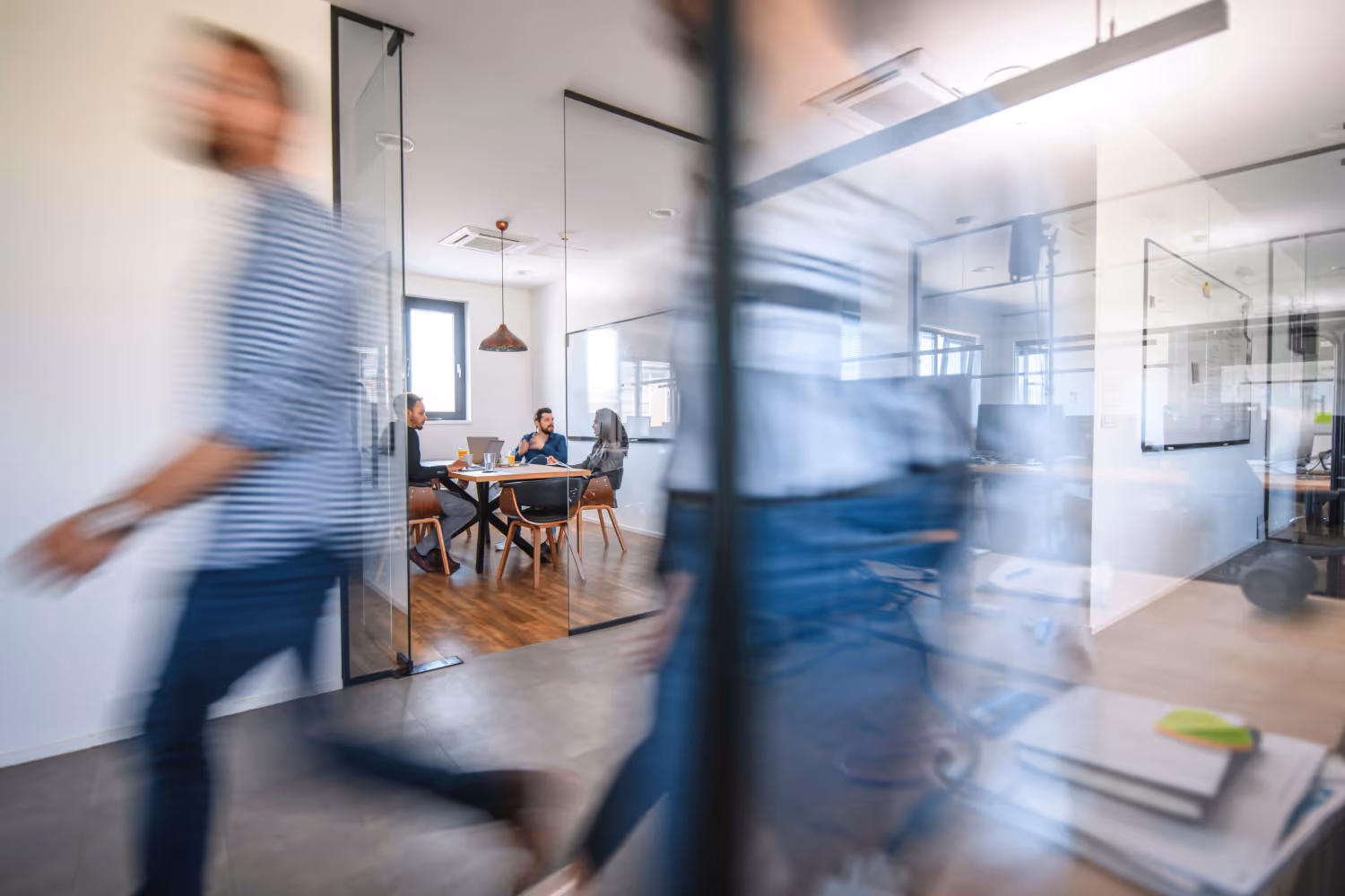 Three people sitting at a table in a modern office conference room having a discussion while two blurred figures walk past glass walls.
