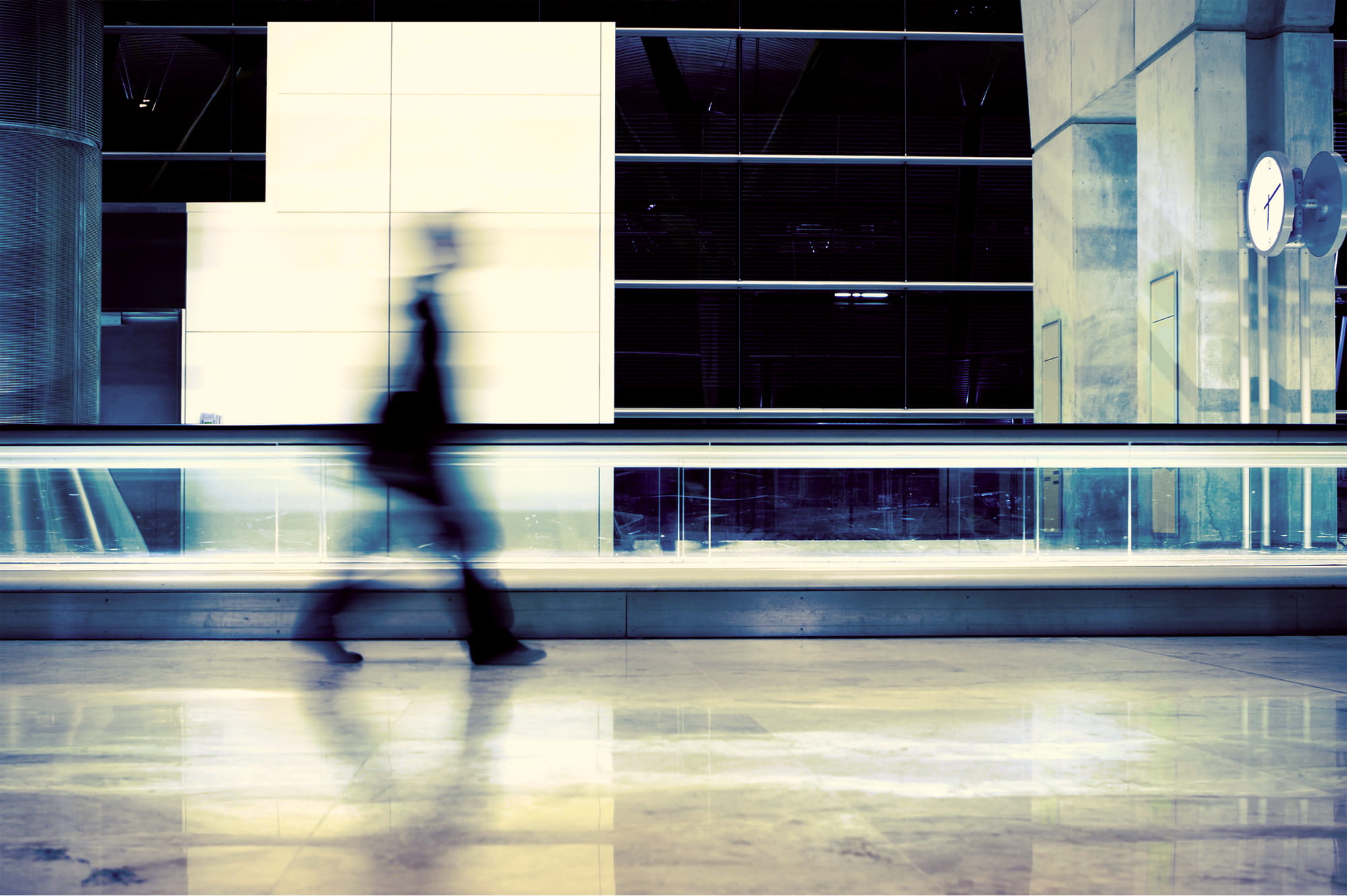 Blurred silhouette of a person walking in a modern, brightly lit indoor space with reflective flooring and a clock on a concrete pillar.