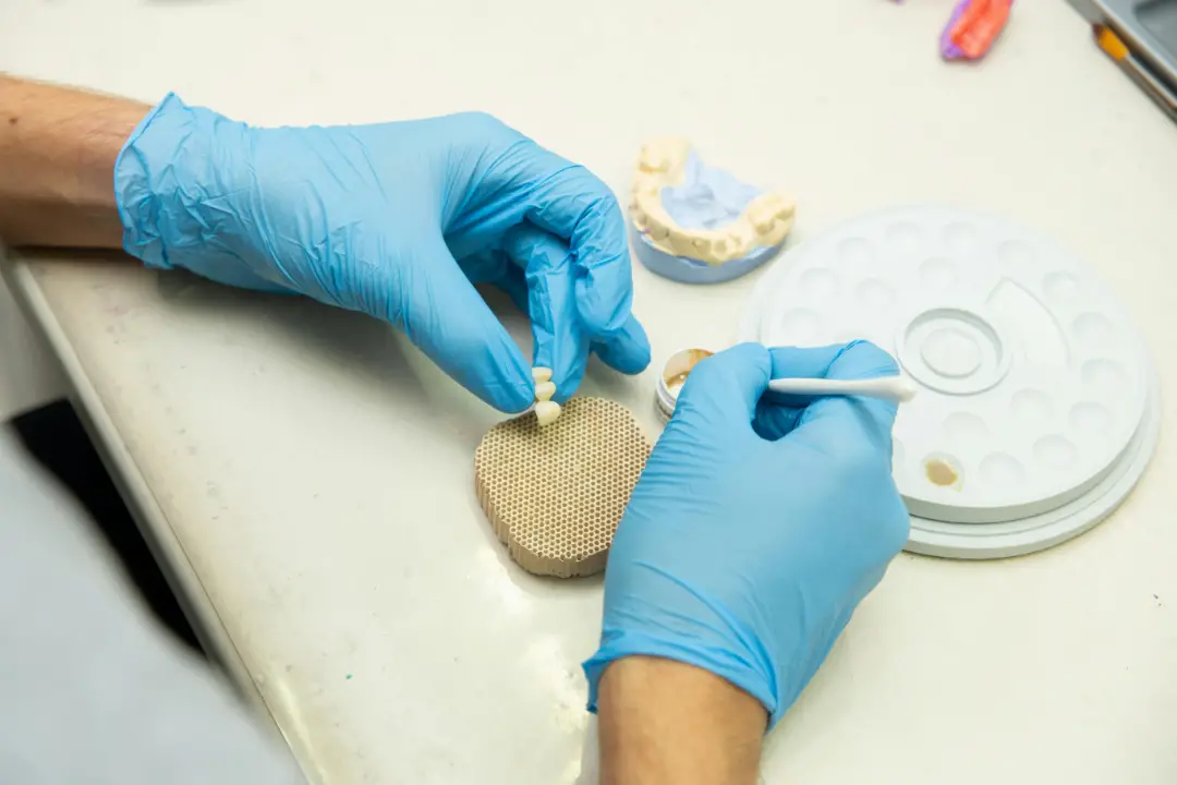 A person wearing blue gloves holds a dental crown in one hand and a cotton swab in the other over a work surface with dental mold and tools.