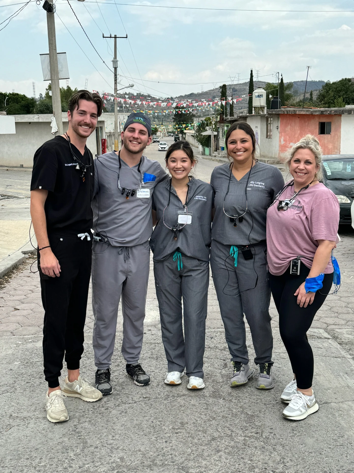 Five smiling dental professionals standing together on a street with colorful triangular flags overhead and houses in the background, four wearing scrubs and one in casual attire with dental loupes around their necks.