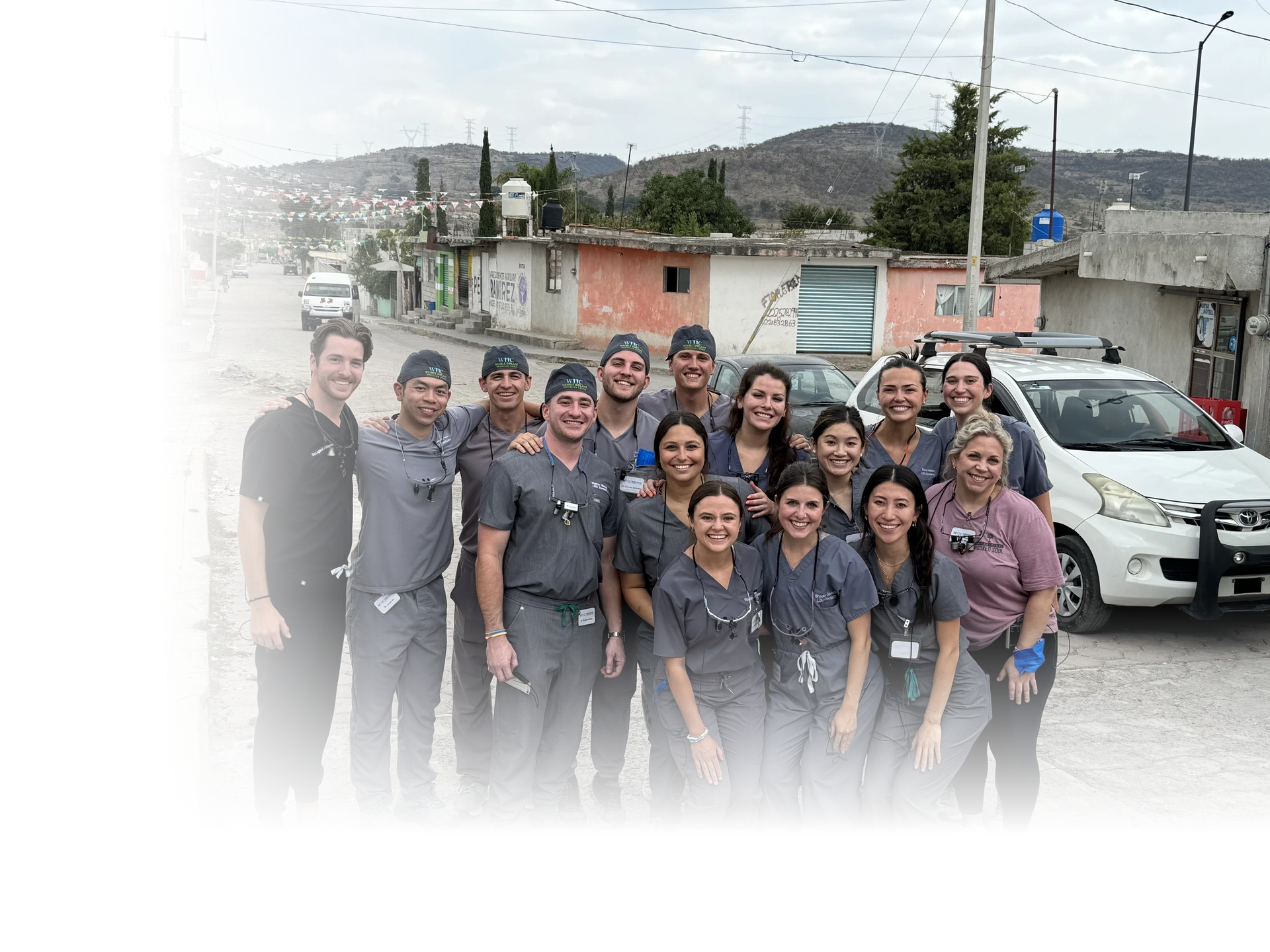 A group of fifteen smiling medical professionals wearing scrubs and dental loupes pose outdoors on a street in front of vehicles and buildings, with hills in the background.
