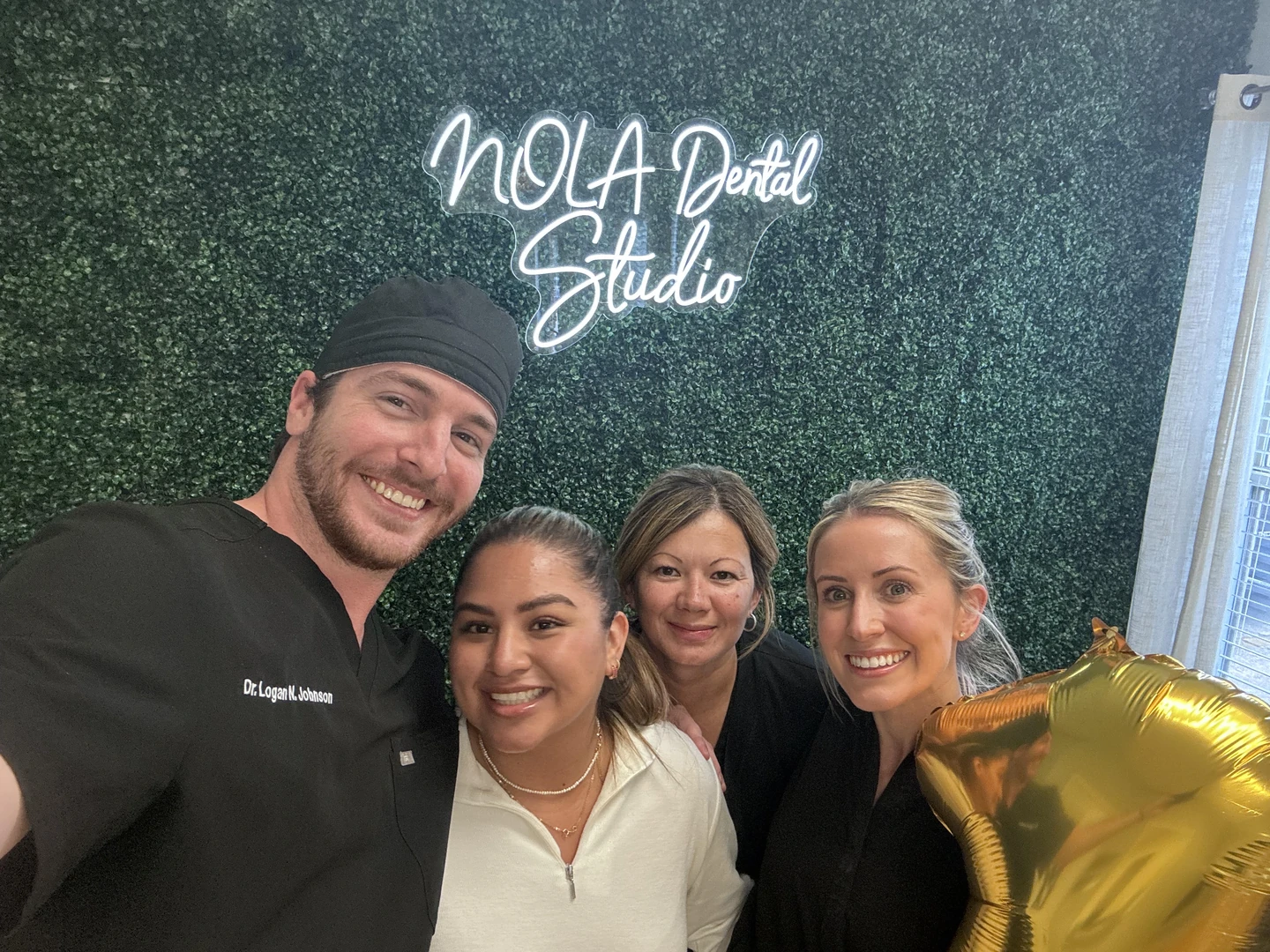 Group of four smiling dental professionals posing for a selfie in front of a green plant wall with a neon sign reading 'NOLA Dental Studio,' one person holding a gold star-shaped balloon.