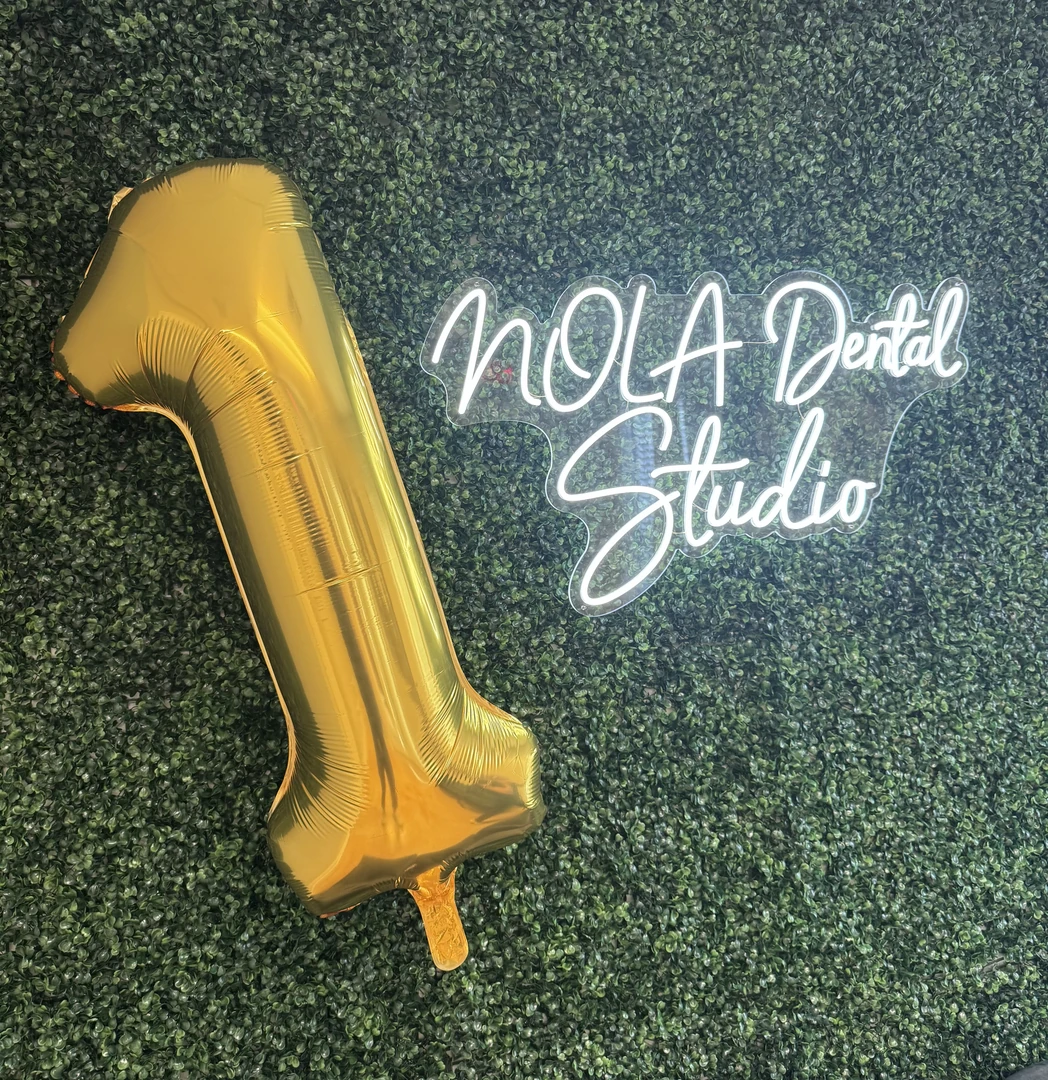 Large gold foil balloon in the shape of the number 1 next to a neon sign reading 'NOLA Dental Studio' on a green leafy wall background.