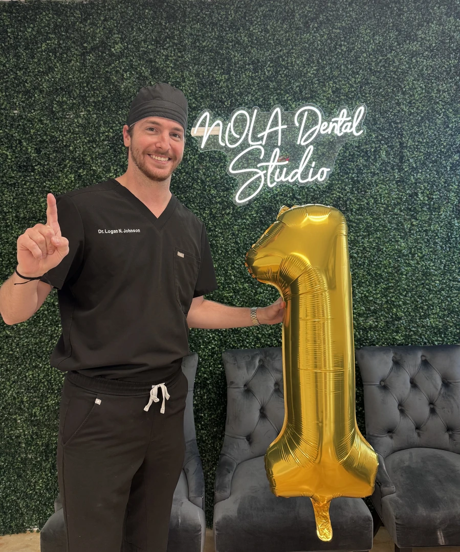 Smiling man in black scrubs with the name tag 'Dr. Logan N. Johnson' holding a large gold balloon shaped like the number 1, standing in front of a green leafy wall with a neon sign that reads 'NOLA Dental Studio'.