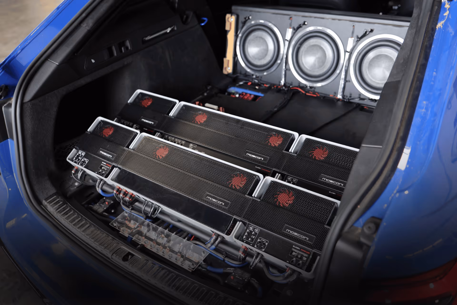 Custom car audio setup with multiple Mosconi amplifiers and subwoofers installed in the trunk of a blue vehicle.
