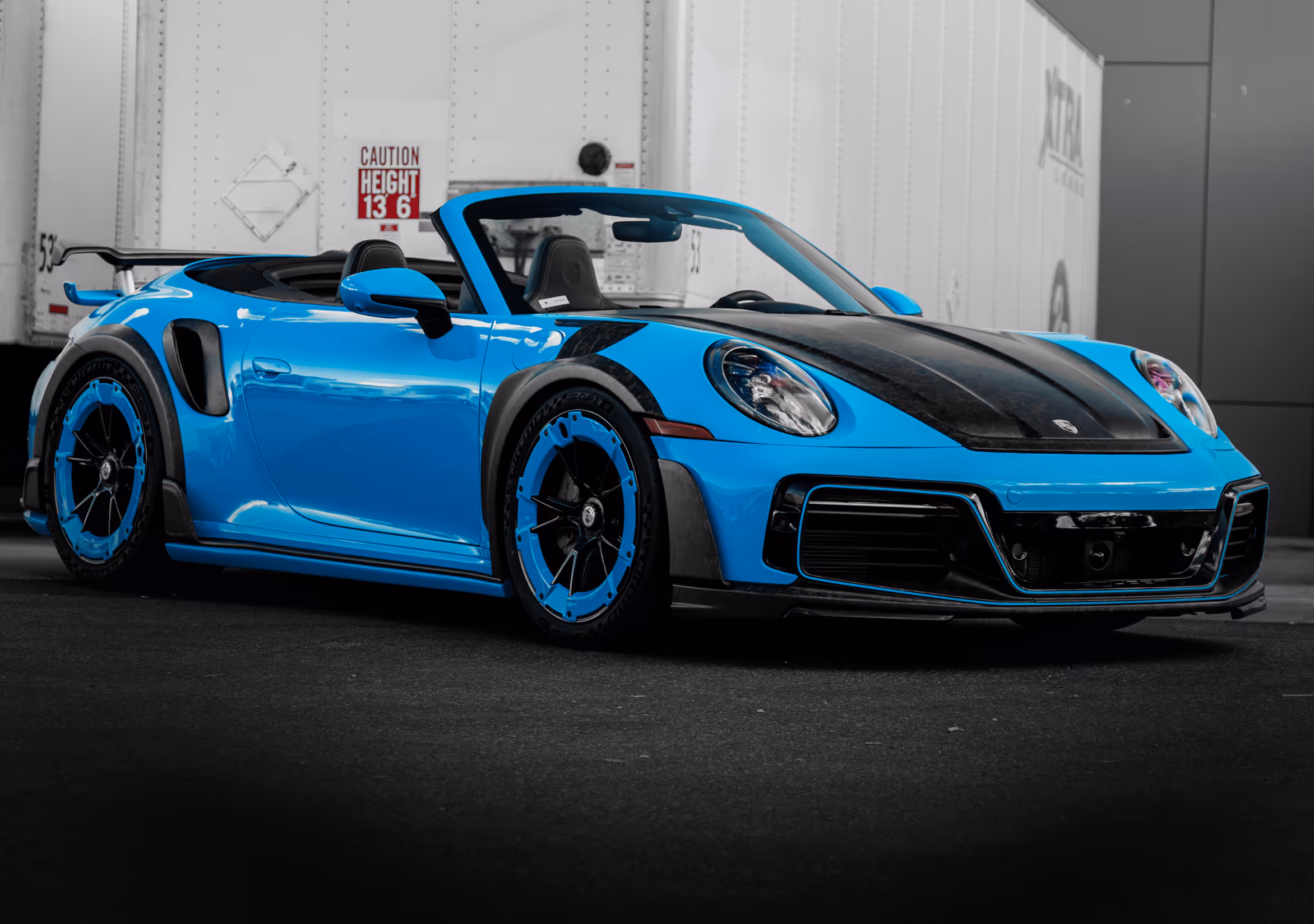 Blue convertible sports car with black accents parked in front of white trailers.
