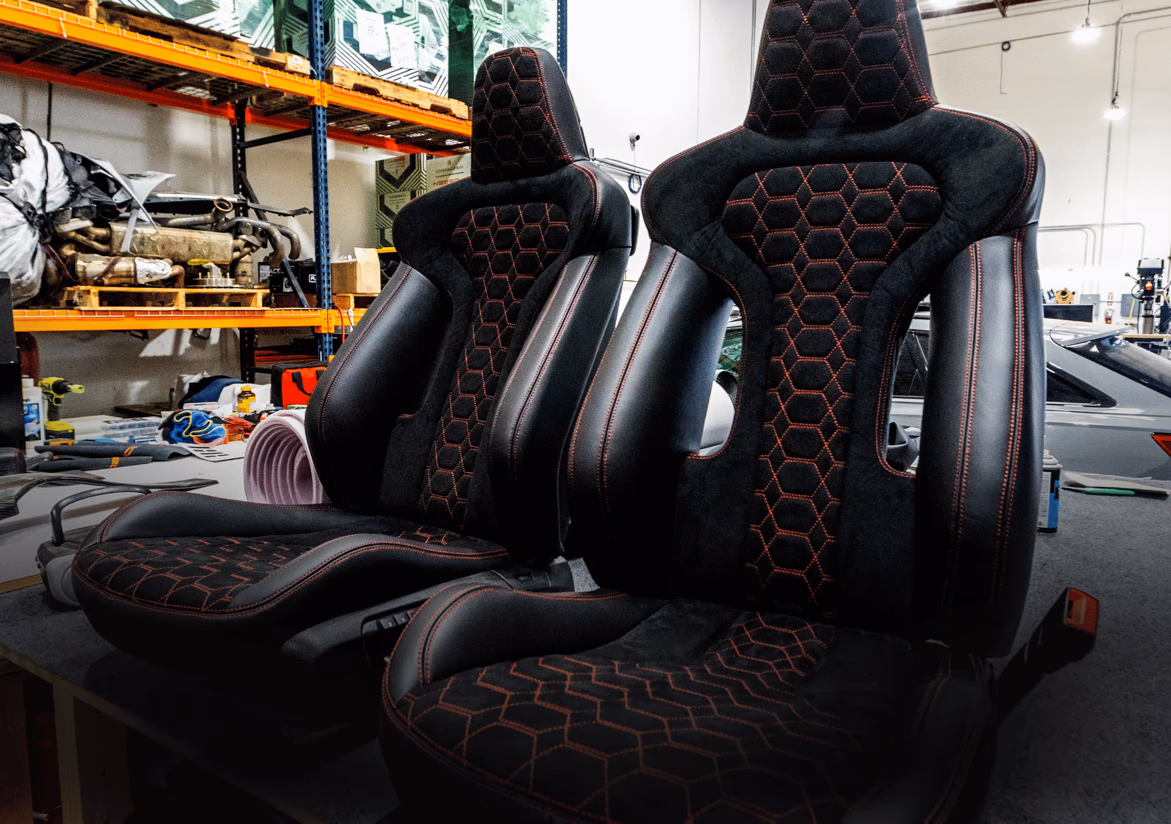 Two black and red racing-style car seats with hexagonal stitching in a workshop setting.