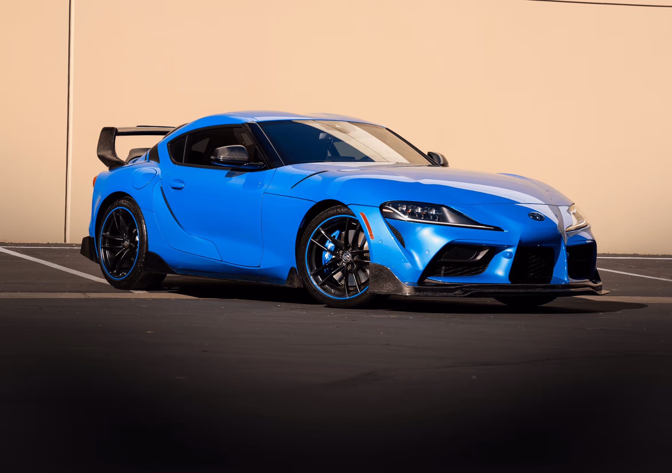 Blue sports car with black rims and rear spoiler parked against a beige wall.