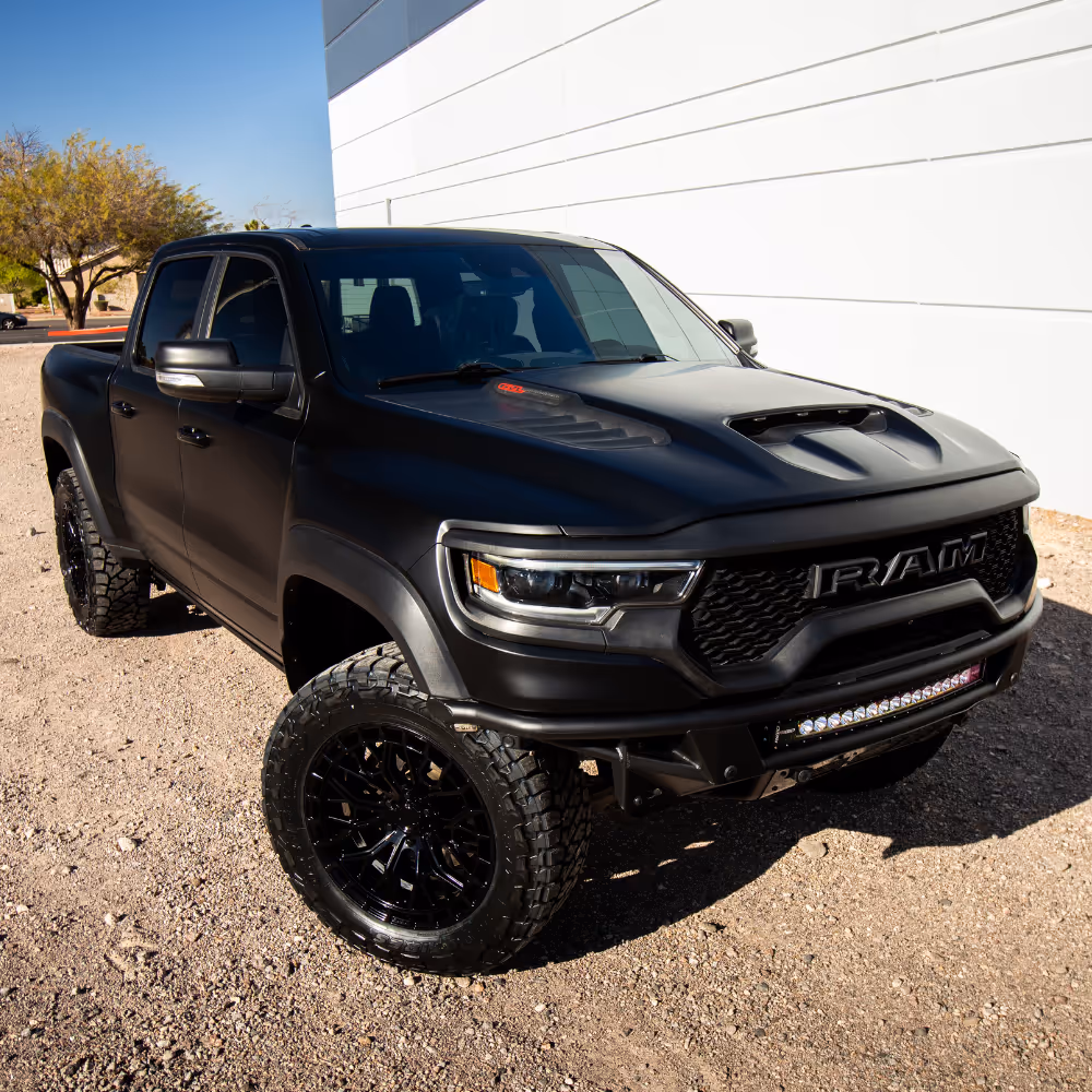 Black RAM pickup truck with off-road tires parked on gravel in front of a white building.