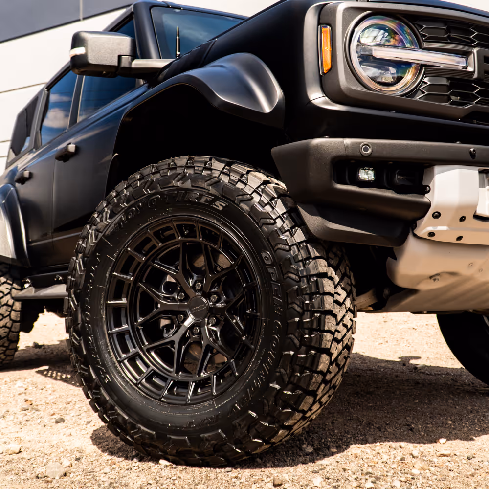 Close-up of a rugged off-road tire and black alloy wheel on a matte black SUV parked on gravel.