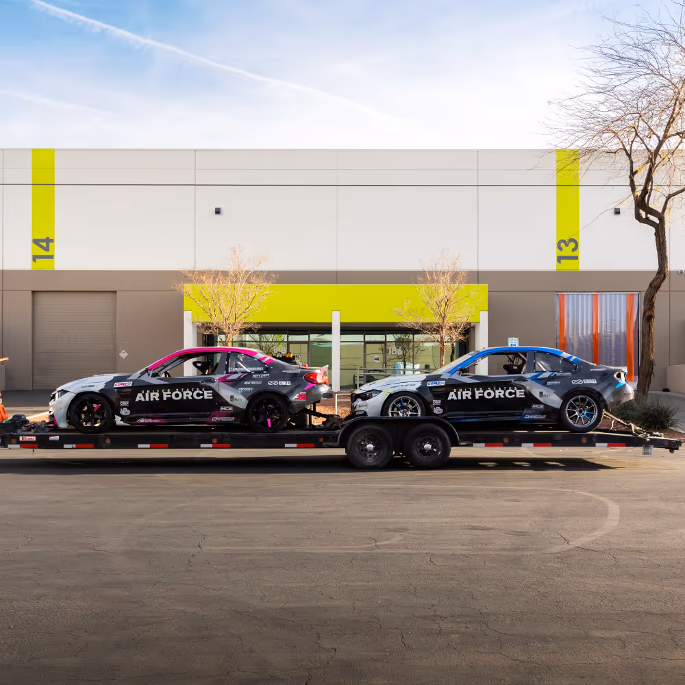 Two black and gray Air Force-branded race cars with pink and blue accents loaded on a trailer in front of a building with green accents and numbered 13 and 14.