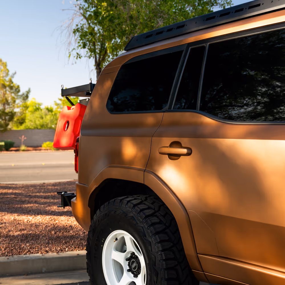 Close-up of the rear side of a bronze SUV with tinted windows, white off-road tires, and a red gas can mounted on the back.