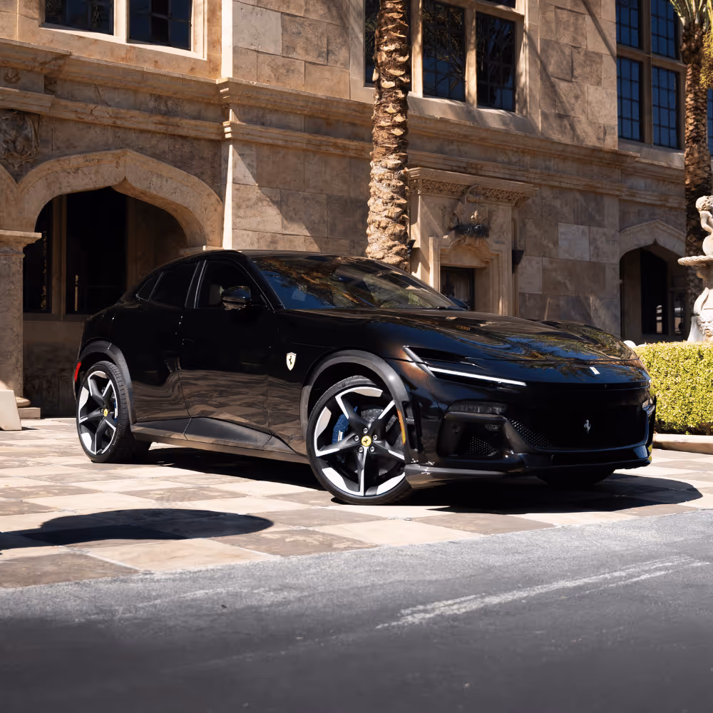 Black Ferrari SUV parked on a stone-tiled driveway in front of a beige stone building with arched windows and palm trees.