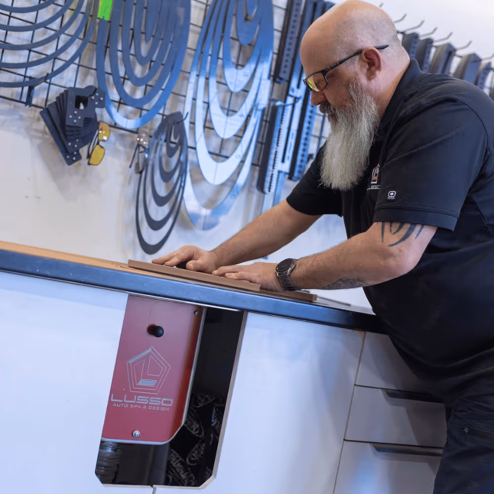 Bearded man with glasses working on a wooden piece at a counter with Lusso Auto Spa & Design branding in the background.