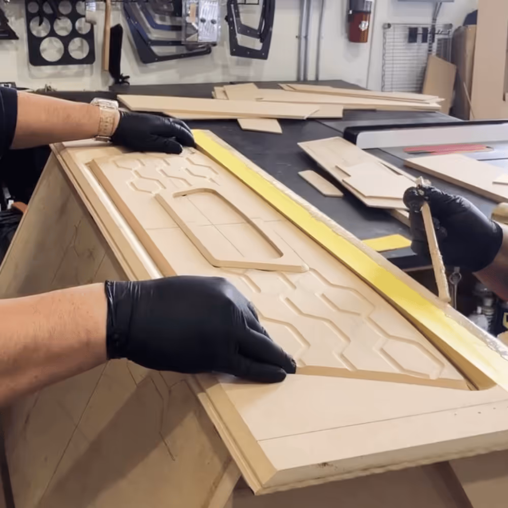 Person wearing black gloves working on a detailed wood panel with geometric patterns in a workshop.