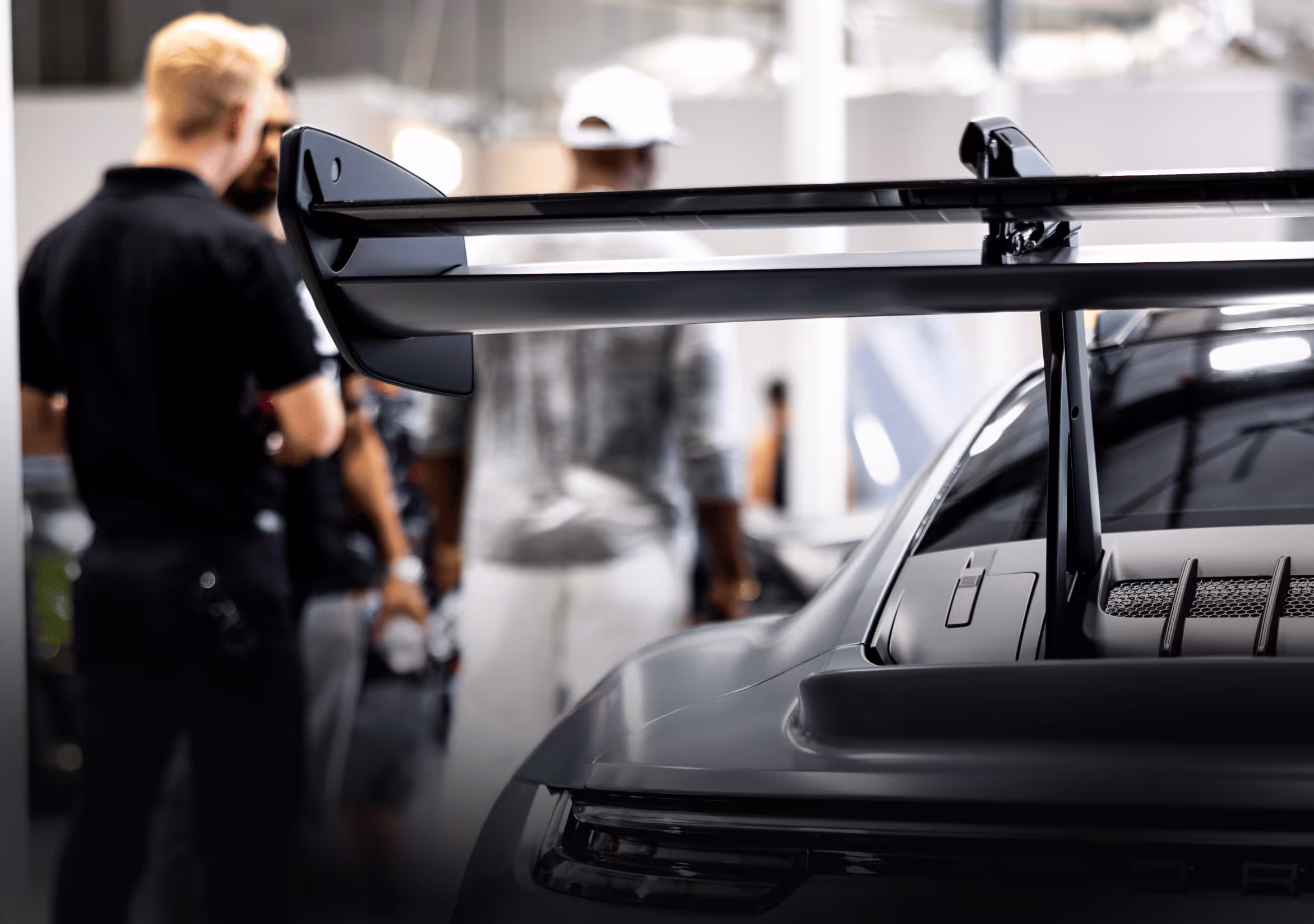 Close-up of a black sports car rear wing with blurred people in the background.