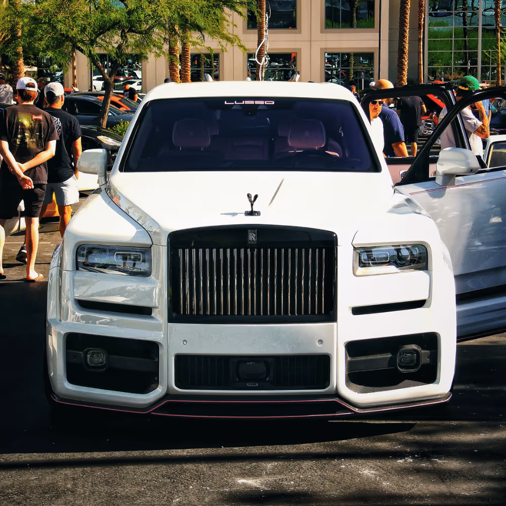 Front view of a white Rolls Royce with a black grille and the Spirit of Ecstasy hood ornament, parked outdoors with palm trees and people in the background.