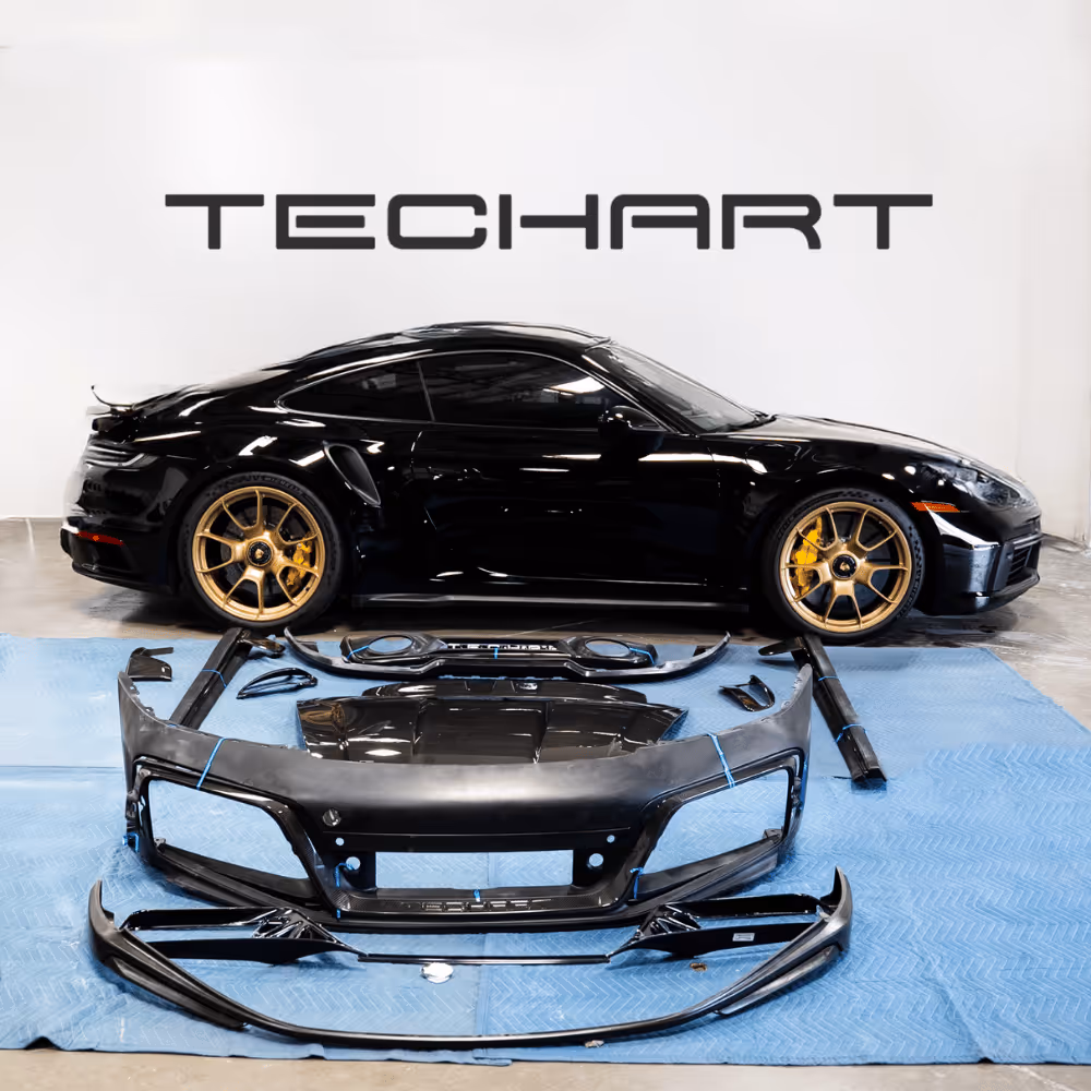 Black TechArt Porsche sports car with gold rims, displayed with disassembled black body kit parts on a blue mat in a garage.