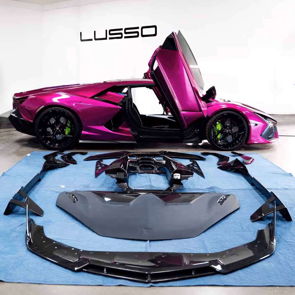 Metallic purple Lamborghini with scissor doors open next to black carbon fiber body kit parts on a blue mat inside a white studio with Lusso logo on the wall.