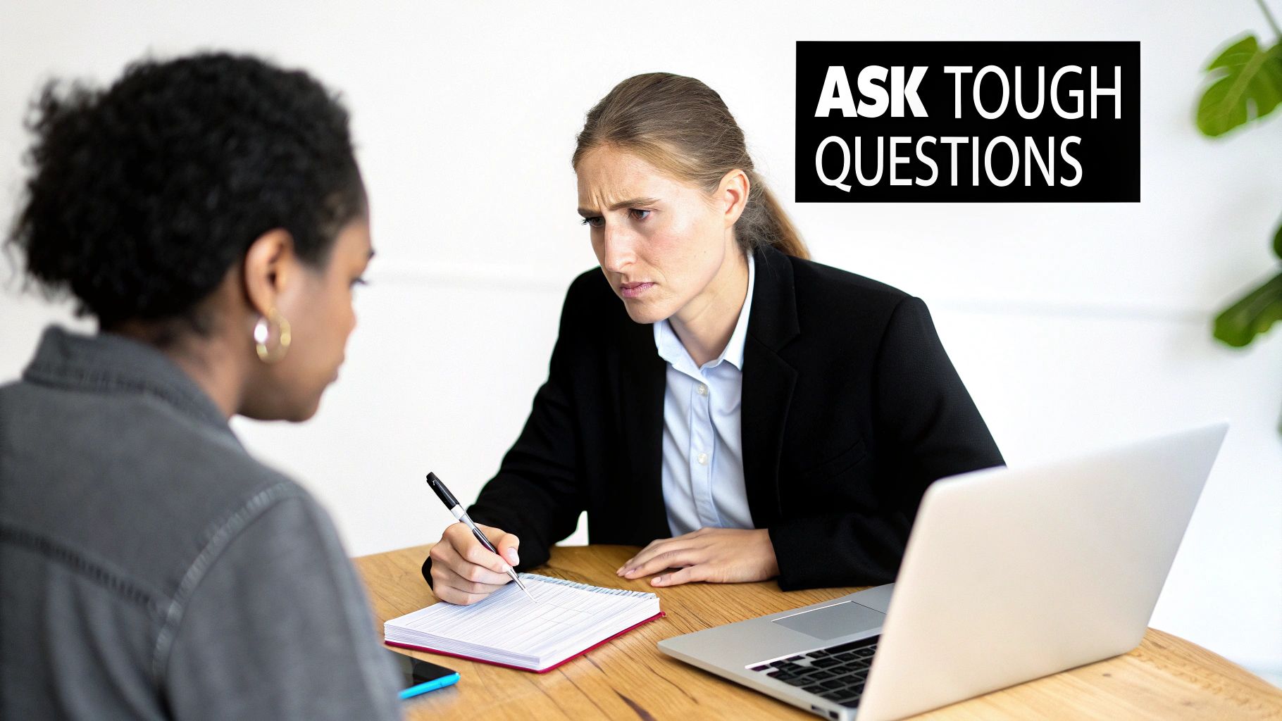 Two professional women engaging in a serious discussion, one takes notes while the other listens.