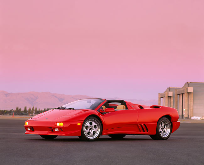 Red convertible sports car parked on tarmac with a pink sky and mountains in the background.