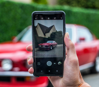 Hand holding a smartphone displaying a photo of a red classic car parked in front of a house, with a blurred red car in the background.