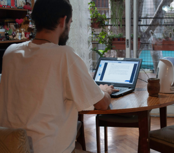 Man in white t-shirt working on a laptop at a wooden table by a window with plants and outdoor view.