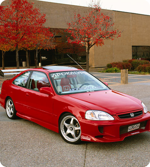 Red modified Honda Civic coupe parked on pavement with autumn trees and building in background.