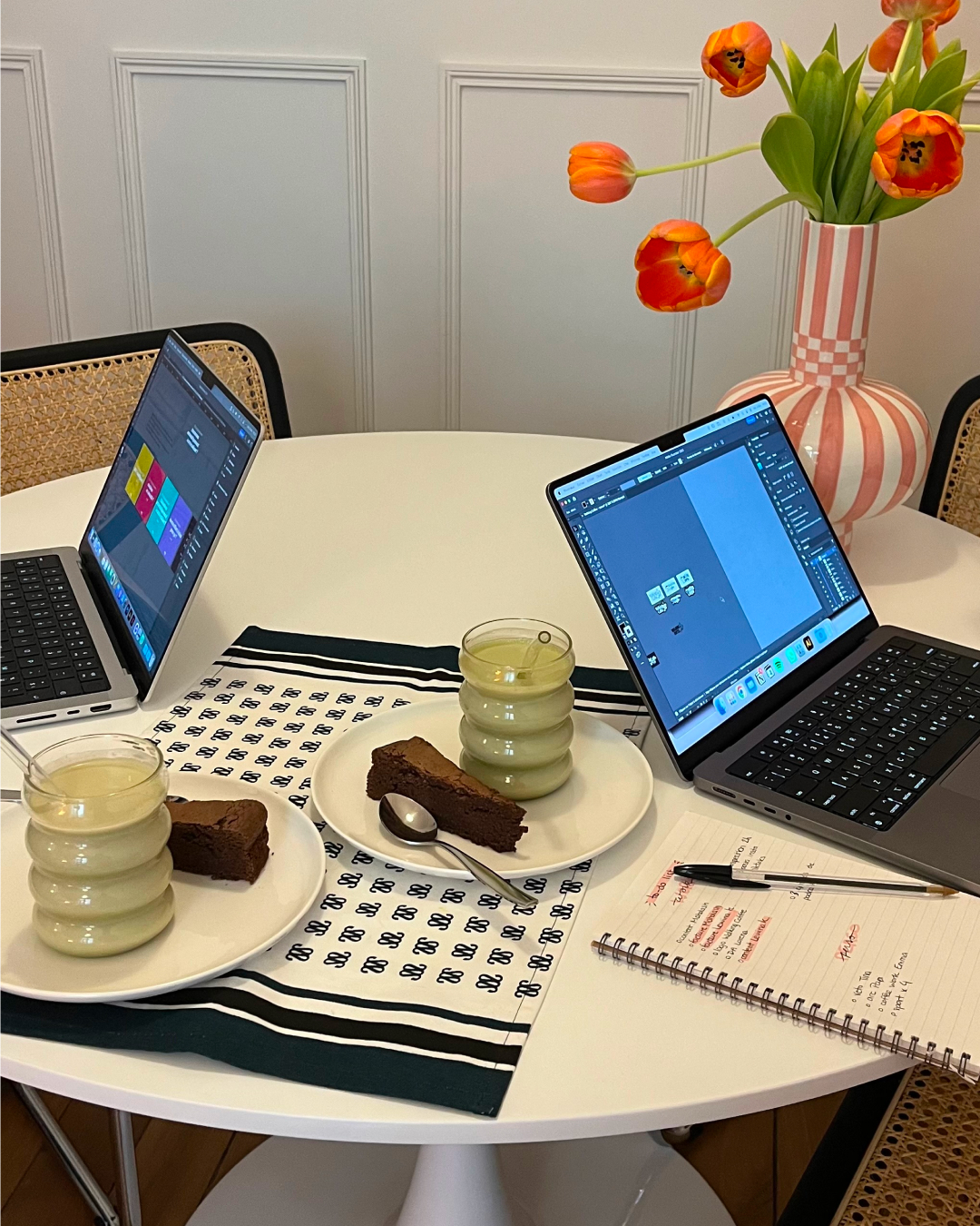 Two laptops on a round white table with plates holding slices of chocolate cake and glasses of green smoothie, a notebook with a pen, and a striped vase with orange tulips.