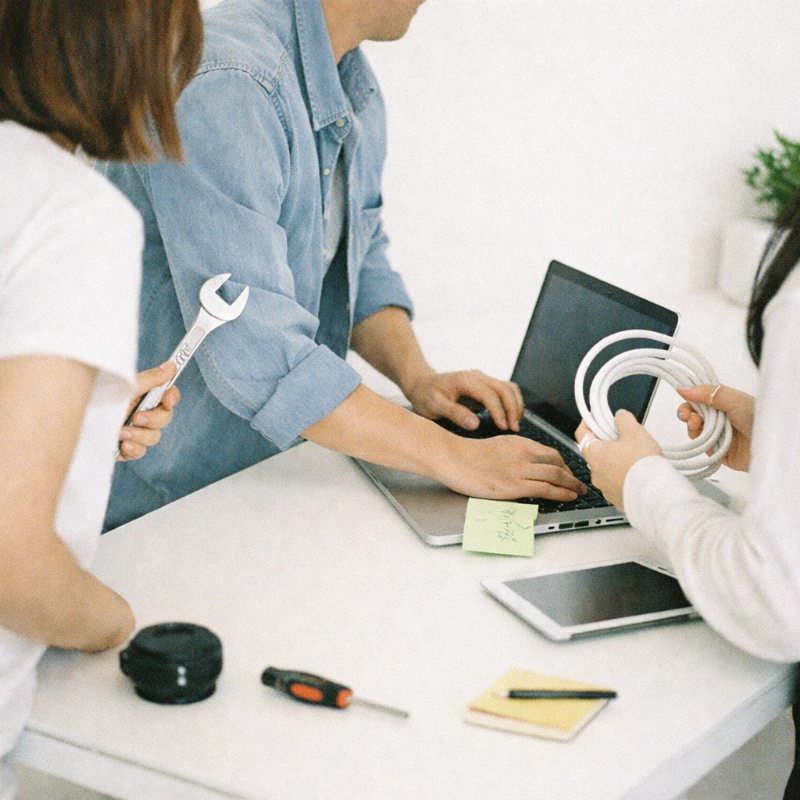 Three people collaborating at a table with a laptop, a coil of cable, and tools including a wrench and screwdriver.