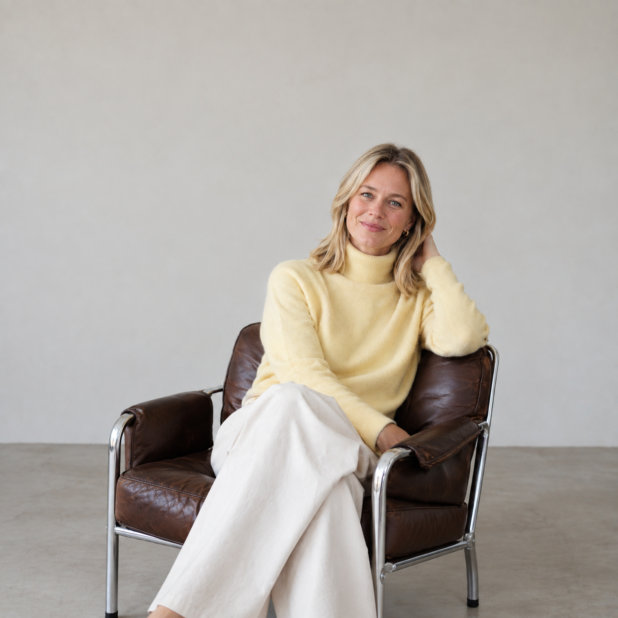 Smiling blonde woman with blue eyes sitting cross-legged on a brown leather chair, wearing a pale yellow turtleneck sweater and white wide-legged pants against a plain light gray background.