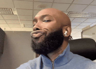 A bald man with a beard wearing a blue zip-up jacket looks sideways with puckered lips, raising his eyebrows in a playful expression, seated indoors with a ceiling and office chair visible.