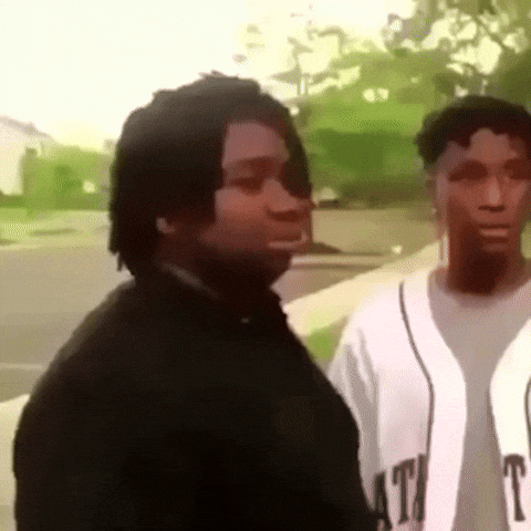 Two young men outdoors, one in a black shirt and the other in a white baseball jersey, standing on a street with greenery in the background; the man in black makes a hand gesture pretending to shoot a finger gun, then makes a surprised facial expression.