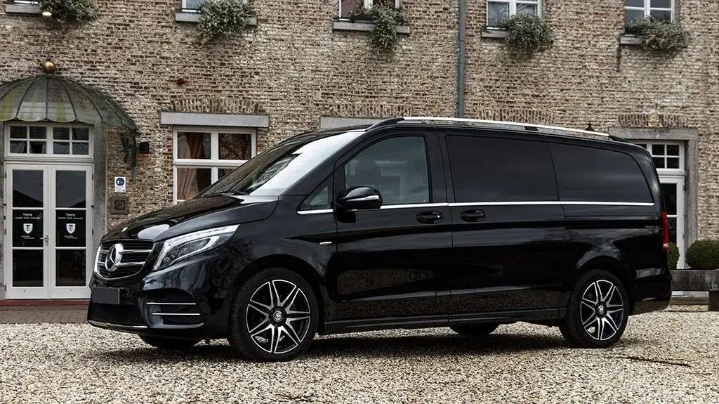 Een luxe, zwarte Mercedes-Benz V-Klasse staat geparkeerd op een grindpad voor een bakstenen gebouw met ramen en deuren tijdens onze privé tour.